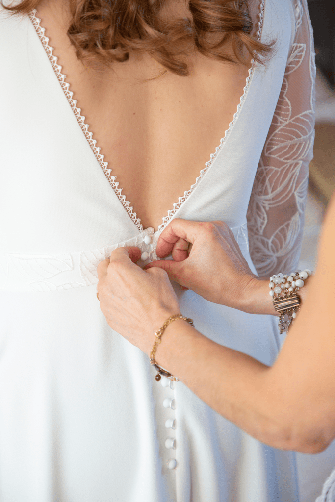 photo de la robe de mariée entrain d'être boutonnée par la mère de la mariée. 
mains avec bijoux
