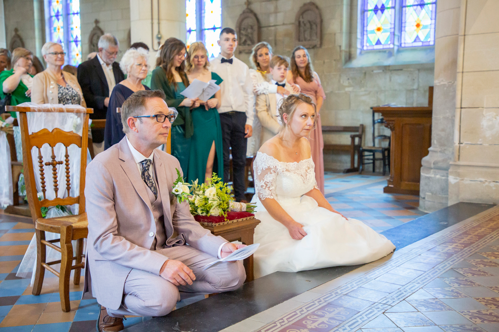 photo mariés à l'église à genoux avec les invités derrière et un bouquet de fleurs